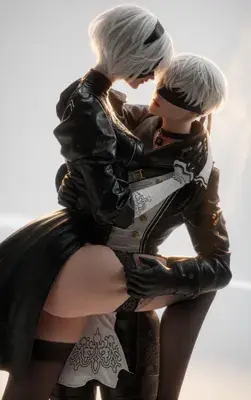 Cosplay photo of two NieR: Automata characters: 2B in a black leather jacket over a thigh-high-slit dress and 9S in a brown-and-white outfit, both with silver hair and blindfolds.