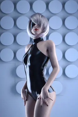 A fan art depiction of 2B (YoRHa No.2 Type B) from NieR: Automata wearing a black latex bodysuit and blindfold, standing against a circular patterned wall.