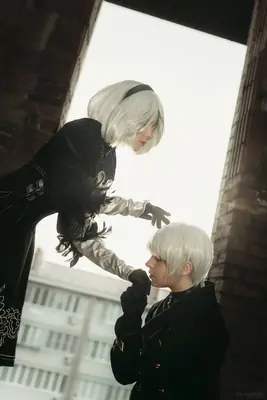 A cosplayer portrays 2B from NieR: Automata, standing in a dramatic pose with another person in an urban setting.