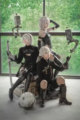 Three cosplayers dressed as 2B, 9S, and A2 from NieR: Automata pose indoors with a green forest backdrop.