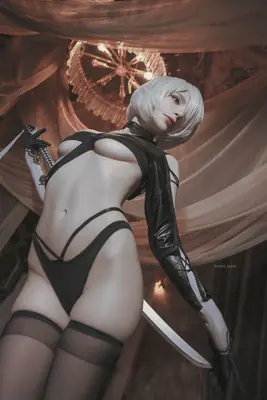 Cosplay of 2B (YoRHa No.2 Type B) from NieR: Automata wearing a black gothic-lolita outfit with a blindfold and sword, posed indoors under draped fabric.