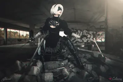A cosplayer portrays 2B from NieR: Automata, wearing a black gothic-lolita outfit with a blindfold, seated amidst rubble in an abandoned industrial building.