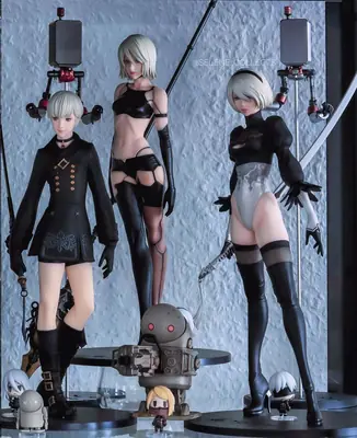 Three 2B (YoRHa No.2 Type B) figures from NieR: Automata are displayed on stands with a small robot and other collectibles in front of a textured wall.