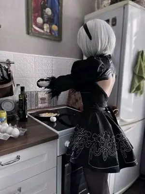 A cosplayer dressed as 2B from NieR: Automata stands in a kitchen preparing food, wearing a black gothic-lolita dress with white embroidery and a blindfold.