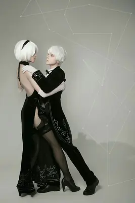 A fan art illustration of 2B and A2 from NieR: Automata, featuring two characters in black gothic-lolita outfits with white hair and blindfolds, embracing against a geometric background.