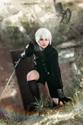A cosplayer portrays 2B (YoRHa No.2 Type B) from NieR: Automata, wearing a black dress and holding a sword in a post-apocalyptic setting.