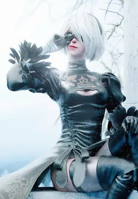 A cosplayer portrays 2B (YoRHa No.2 Type B) from NieR: Automata, wearing a black gothic-lolita outfit with a blindfold and gloves in a snowy outdoor setting.