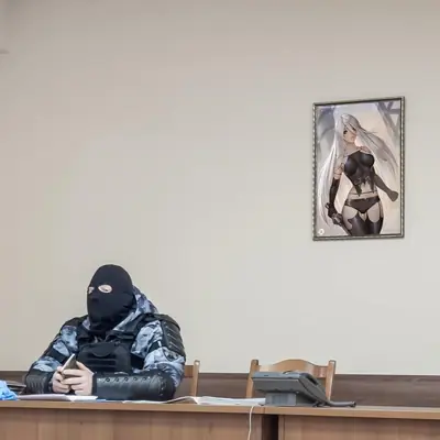 A framed digital illustration of 2B from NieR: Automata hanging on a wall above a person in tactical gear.